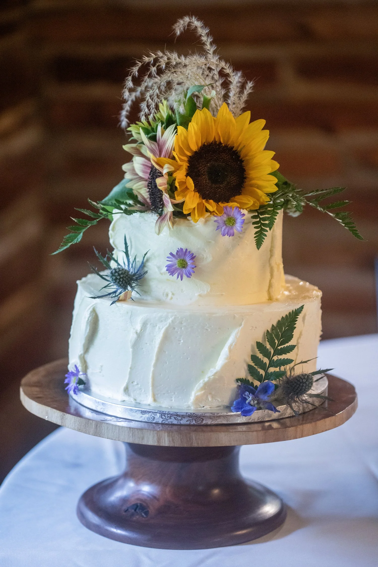 A two-tired round cake with white buttercream frosting sits on a wooden cake stand on a table with a white tablecloth in front of a brick wall. The cake is decorated on the sides with small purple flowers and fern leaves. The top is a sunflower with 