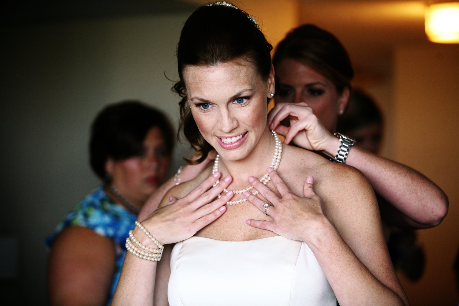 A bride with red hair and a white strapless wedding dress holds the pearls at her collarbone as she is assisted in putting it on by two women behind her. She has a huge smile on her face.