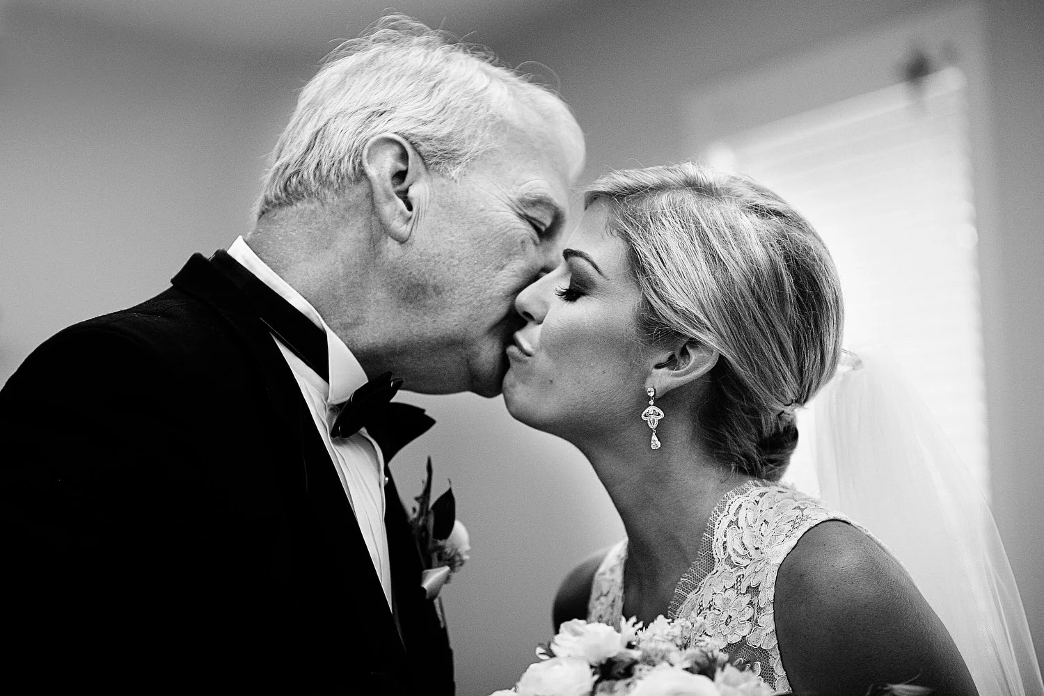 Black and white photo of a bride and her father simultaneously kissing each other on the cheek before her wedding ceremony. They are both close in the frame. She has on a lacy white dress and dangly earrings. Her hair is pulled into a loose bun with 
