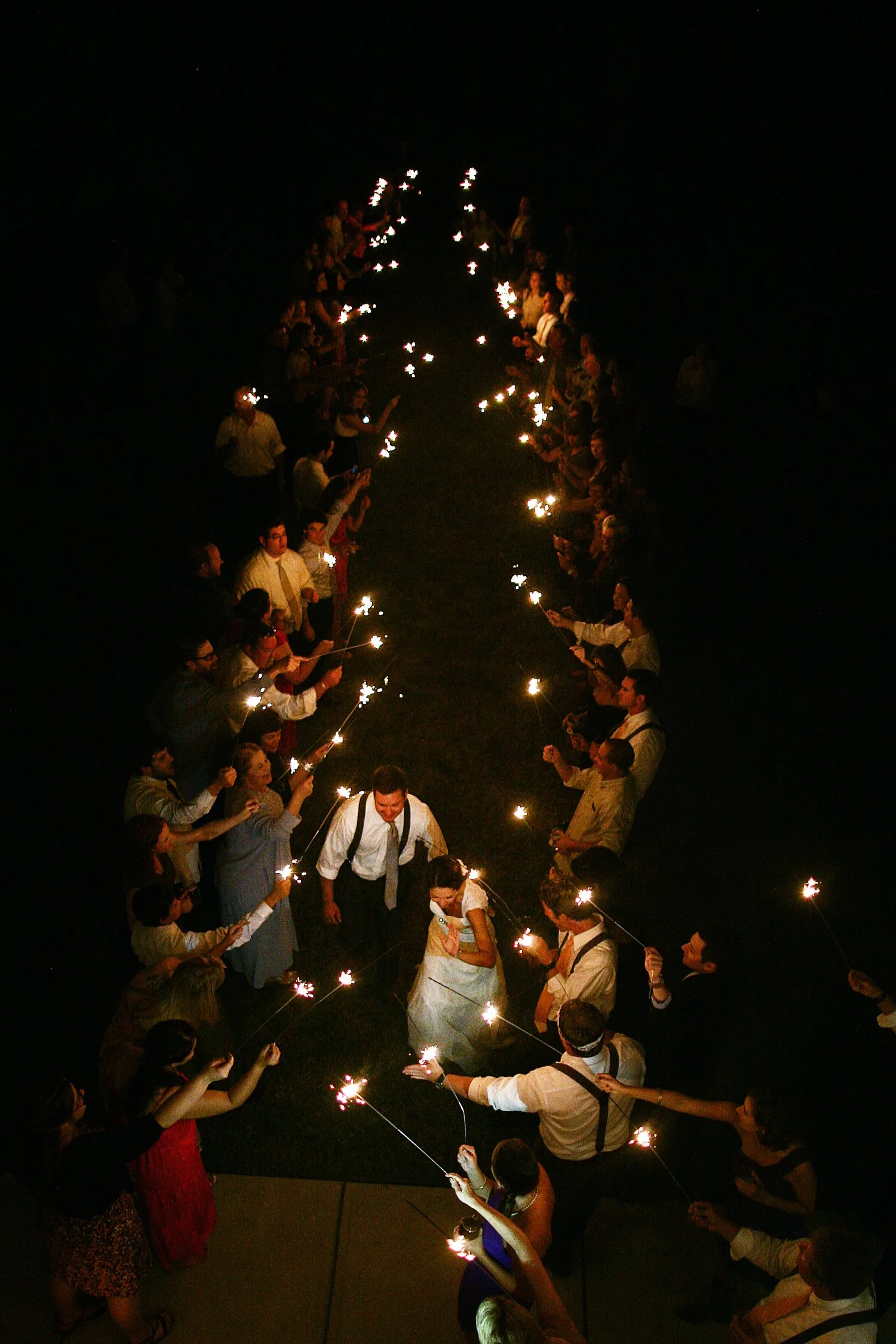 Photo taken from above. A bride and groom exit their wedding reception down a grassy path lined by people holding sparklers down both sides. 