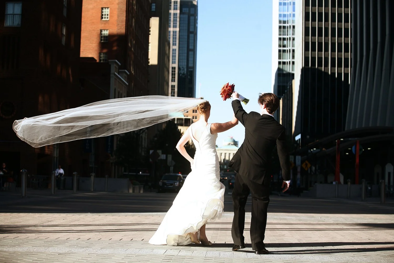 A bride and groom walk down a city street in Raleigh. She is holding her white wedding dress at her waist with her left hand while she holds her right hand in the air clutching his and a bouquet of flowers. Her floor-length veil is blowing out to the