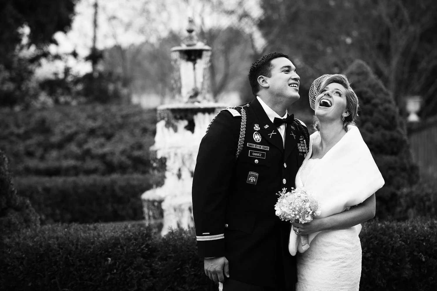 Black and white photo of a bride and groom celebrating outdoors, with the groom in a military uniform and the bride in a wedding dress with a white fur stole and birdcage veil holding a bouquet, both smiling and looking joyful. There is a fountain.