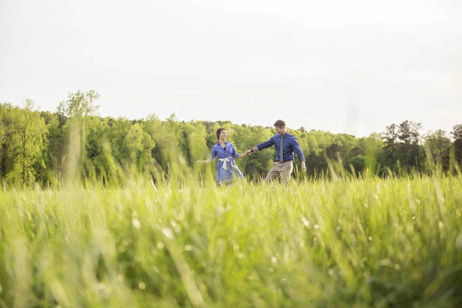 Low angle photo from a distance. The green grassy field is shown in the foreground. In the far distance a couple is seen holding hands and walking. She has on a denim blue dress with a white belt. He has on a blue dress shirt and khakis. 