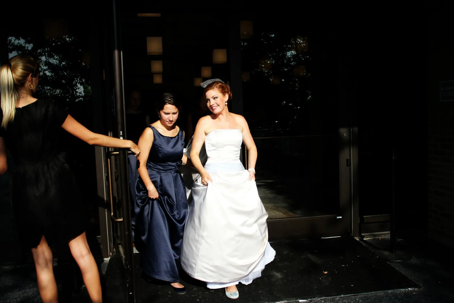 A bride in a white wedding dress standing outside a building with two women in dark blue dresses, one holding the door open. The bride is in stunning light and smiling big.