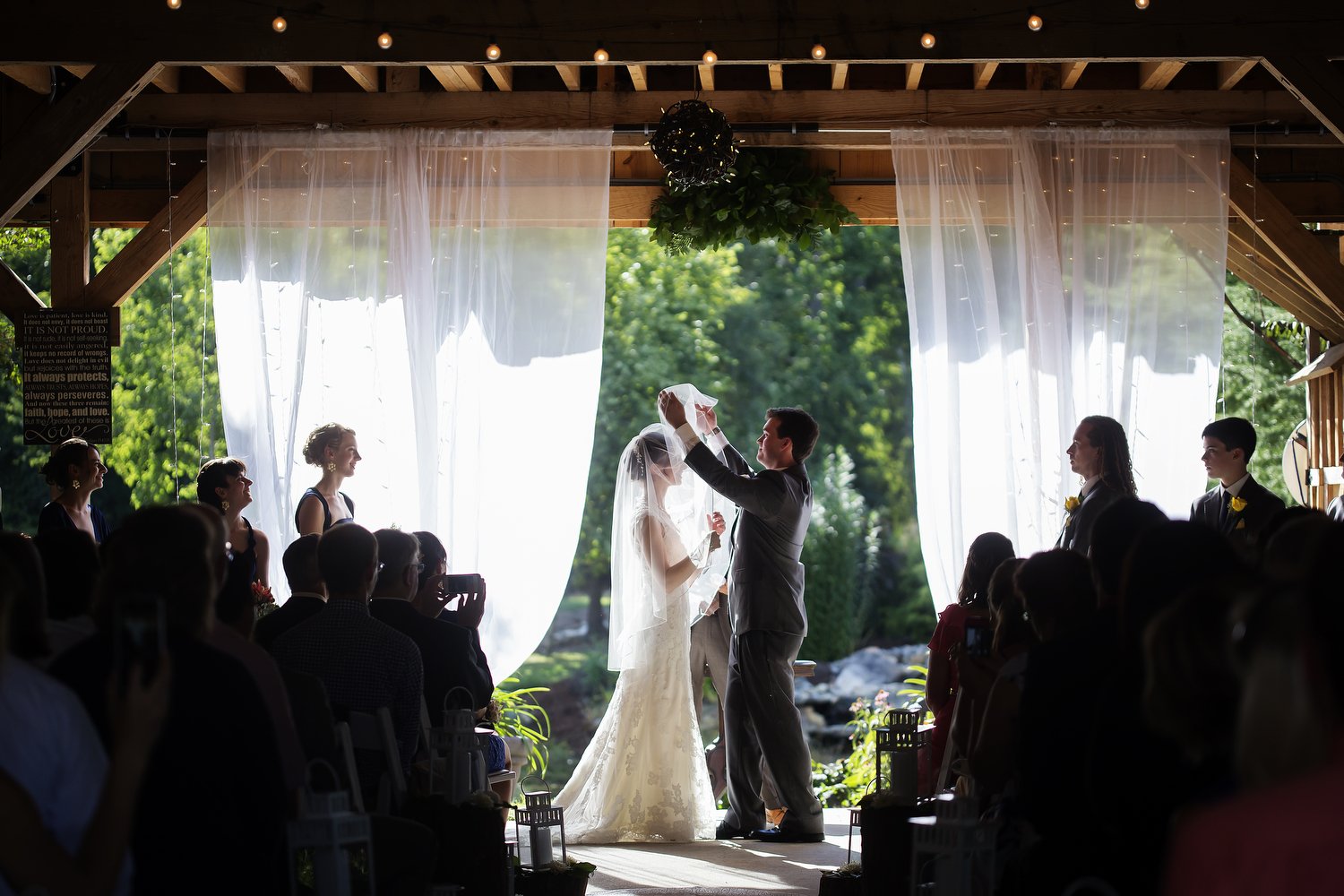 A couple getting married during an outdoor wedding ceremony. The groom lifts the bride's veil, surrounded by guests in a decorated wooden pavilion with sheer curtains and string lights.  Her wedding dress and veil are illuminated by light behind her.