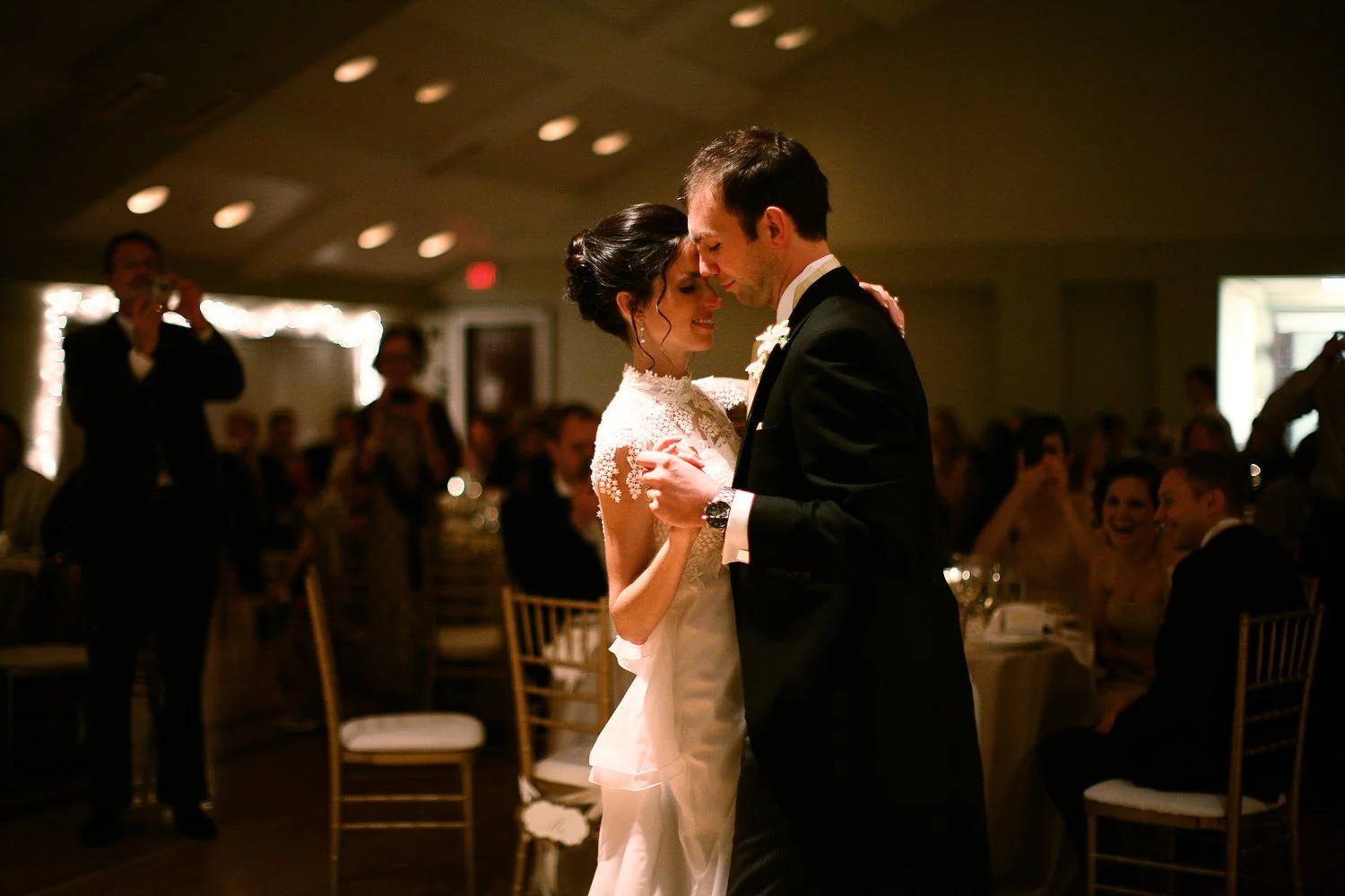 A bride and groom share their first dance at their wedding reception in a dimly lit room, surrounded by seated guests watching and smiling. She is wearing a vintage white dress and he is in a black tuxedo.
