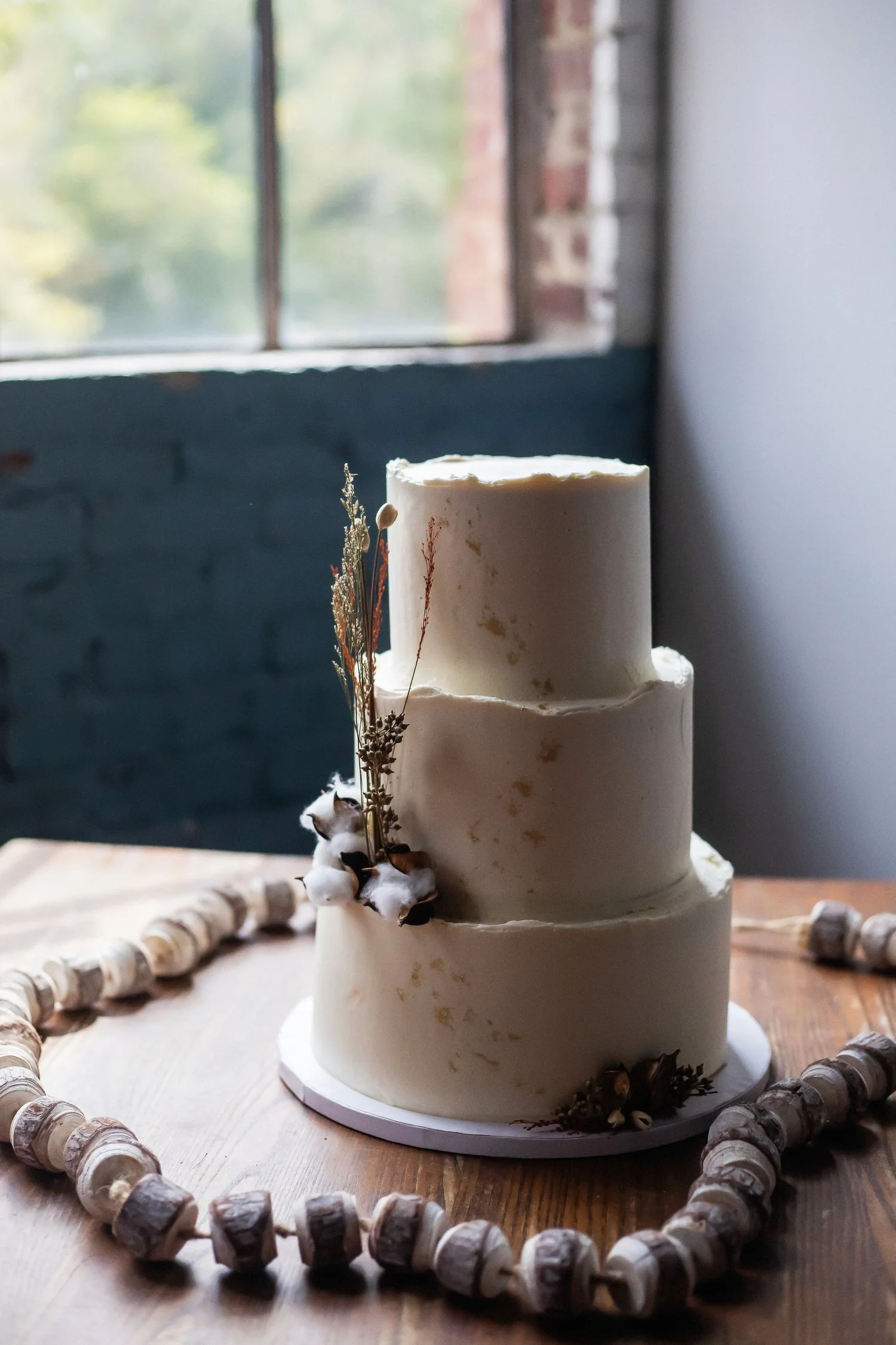 Vintage-styled three tier round cake sits on an old wooden table surface. There are small flecks of gold here and there on the white frosted surface. The bottom tier has some cotton and wheat grain like decorations sticking out of it. A strand of cot