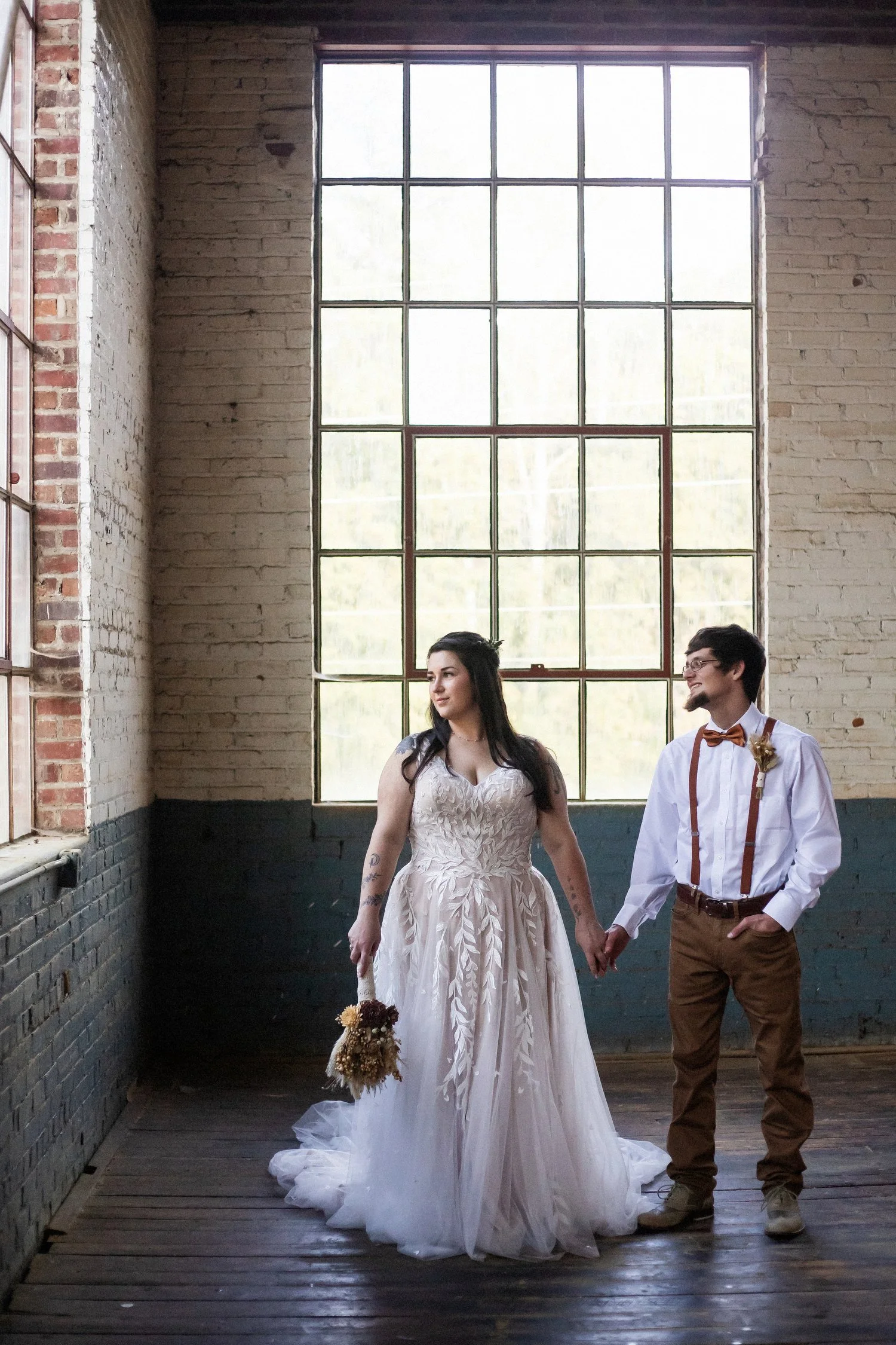A bride and groom holding hands in a rustic indoor setting with brick walls and large windows, the bride in a white gown holding a bouquet and the groom in a white shirt with suspenders and bow tie.