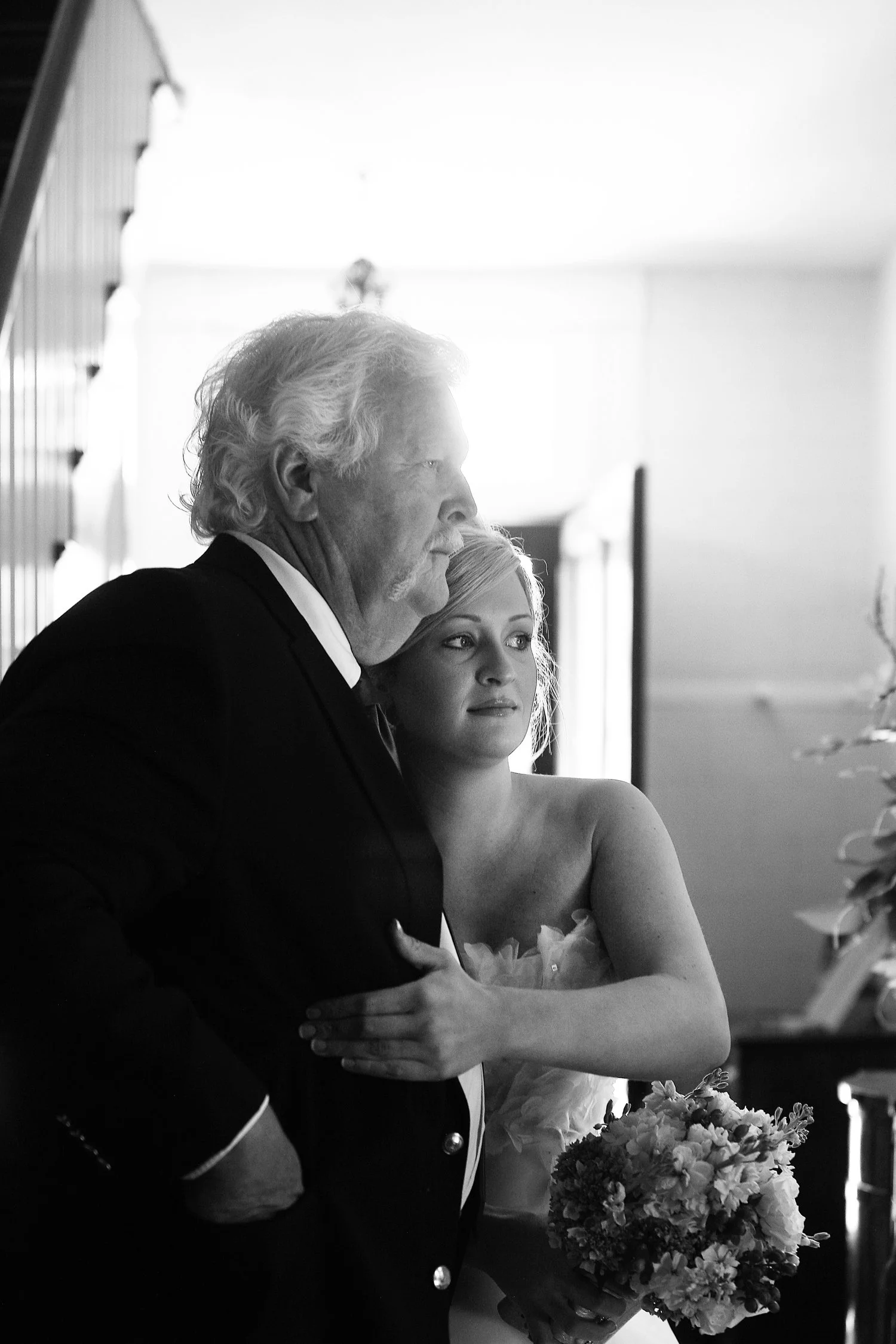 Black and white photo of an elderly man in a tuxedo and a young woman in a wedding dress standing close together, with the woman holding a bouquet of flowers, in a softly lit interior.