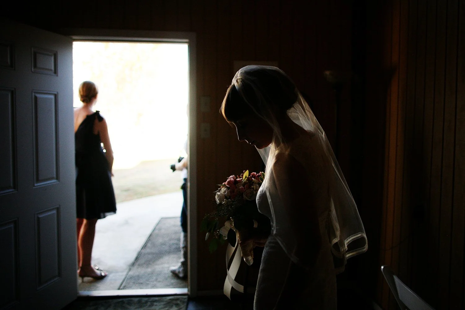 A bride holding a bouquet of flowers, standing in a dark room, with sunlight coming from outside, while a woman stands near the open door. The bride's profile is silhouetted by the gorgeous light coming inside the room from the open door.
