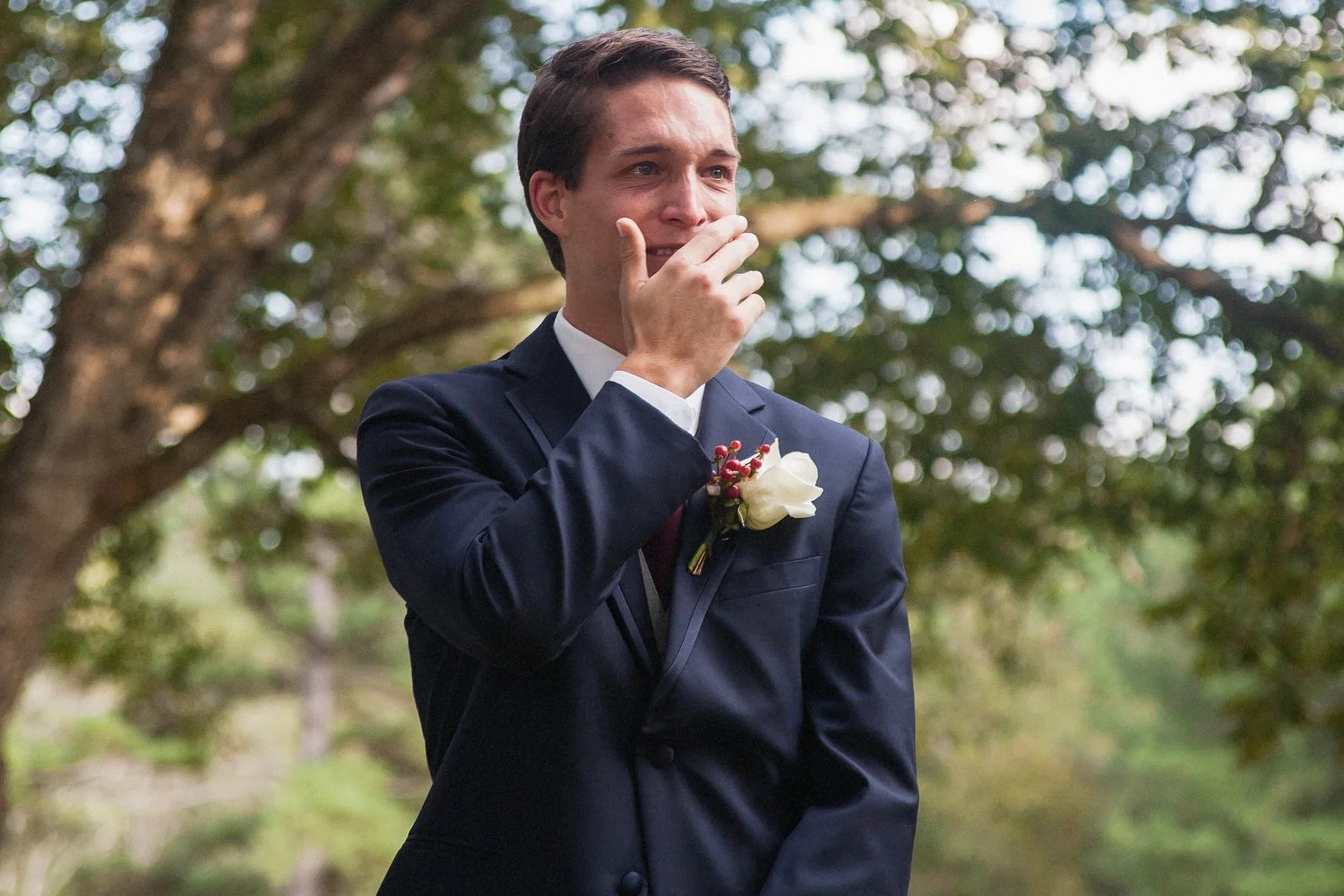 A man in a navy blue suit with a boutonniere, standing outdoors with trees in the background, appears emotional and has his hand covering his mouth as he catches a glimpse of his bride for the first time.
