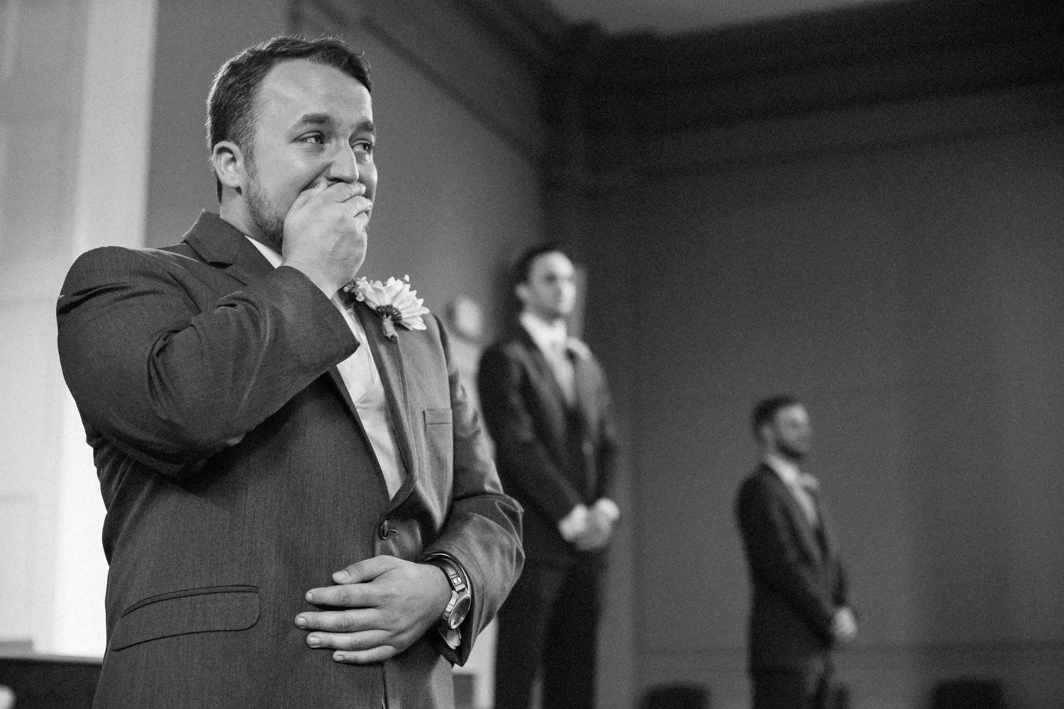 Black and white photo of a groom in a suit covering his mouth with one hand and blinking back tears as he sees his bride for the first time. He is standing in front of two other men also in suits.