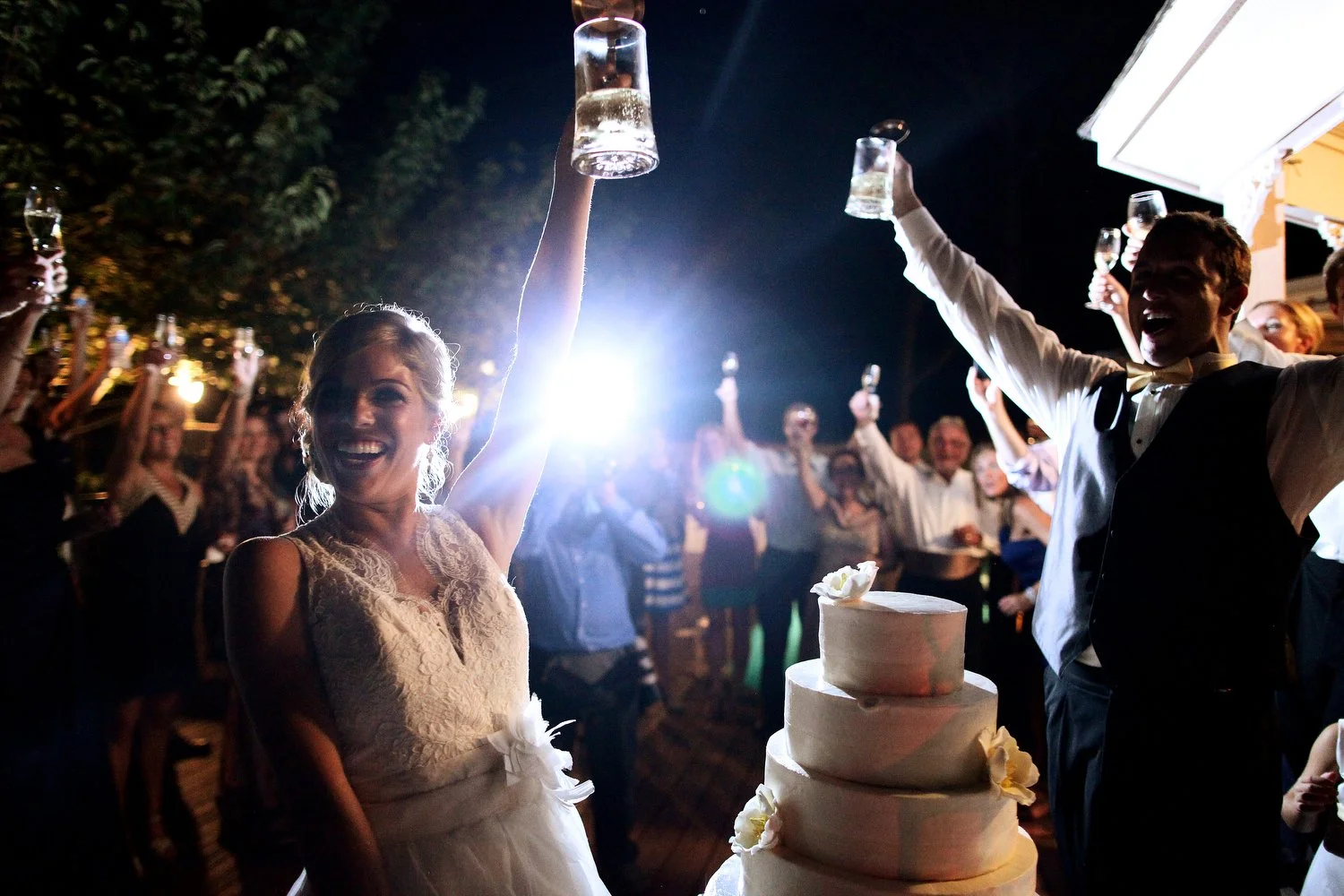 Bride and groom celebrating with champagne glasses at wedding reception, surrounded by guests, outdoors at night, wedding cake in front. Their glasses are held high in the air illuminated by a camera flash.