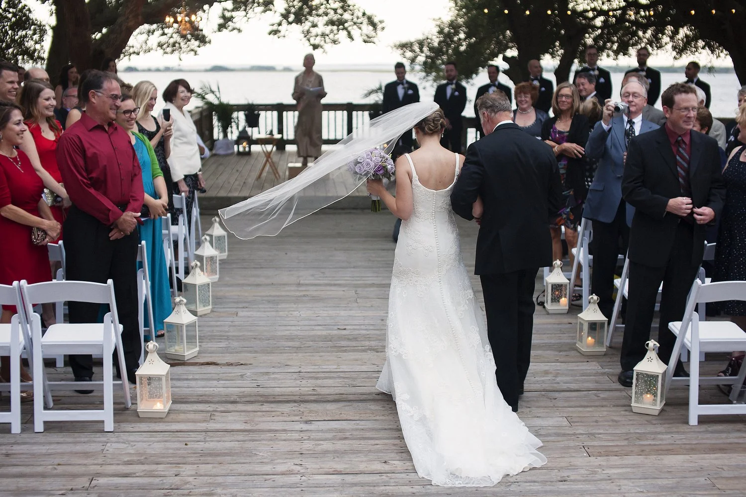 Bride walking down the aisle with her father at an outdoor wedding ceremony by the water, surrounded by guests and lanterns. Her long veil is blowing out to the left beside her as they walk down the wooden dock-like floor.
