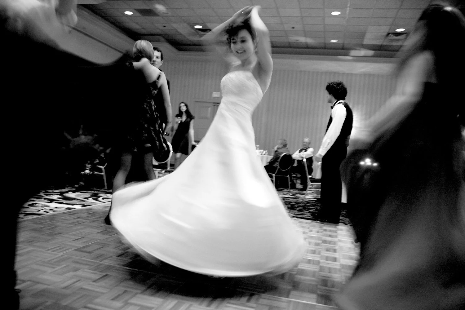 An intentionally blurred image with people off to the sides in the forefront as a bride twirls her strapless white dress in the center of the dance floor with her arms up over her head. The image is black and white.