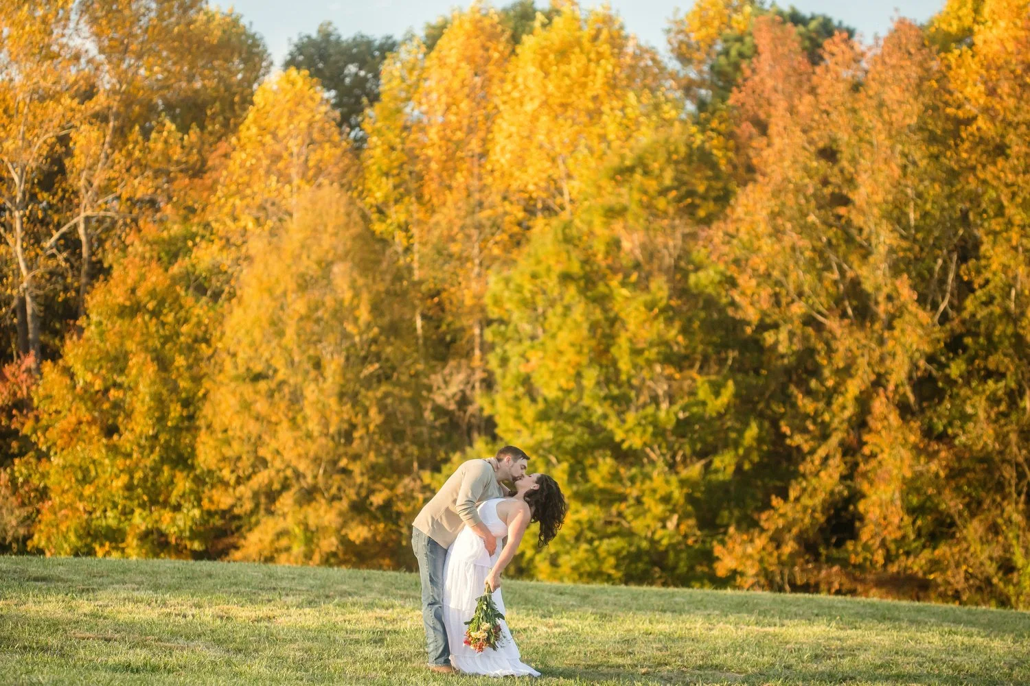 Photo is taken from a distance. In a green field a man dips a woman and kisses her. She leans back wearing a white dress and holding flowers at her side. He has on jeans and a tan long-sleeve shirt. There are huge trees with orange leaves lined up be