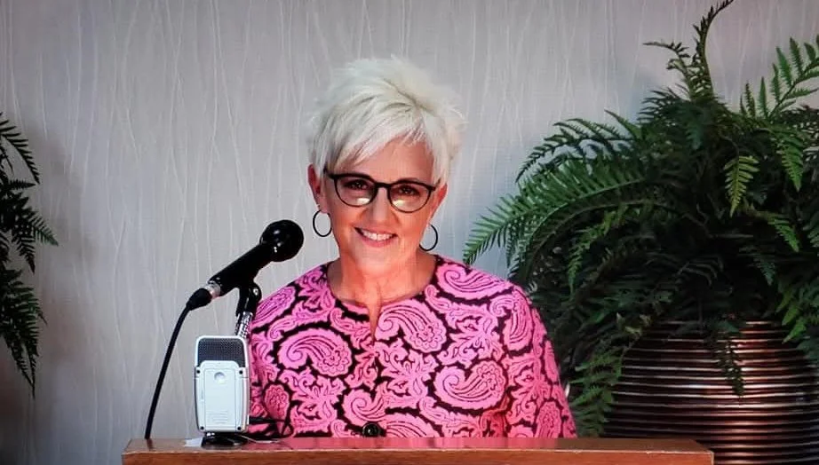 A smiling woman with short, white hair and glasses, wearing a pink and black patterned top, standing behind a podium with a microphone and an electronic device, in front of a large potted plant.