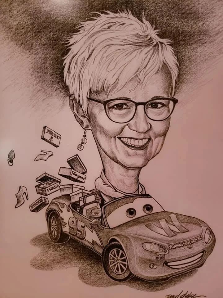 A caricature of a woman with short blond hair and glasses, smiling. She is depicted as a race car driver sitting in a race car with the number 95, labeled "Winning Edge Graphics." The car has anthropomorphic eyes and a wide smile. Various items, including shoe boxes, lipstick, and jewelry, are flying out of the car's trunk.