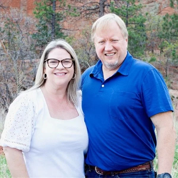 A smiling woman with glasses and long blonde hair standing outdoors next to a smiling man with short blonde hair in a blue polo shirt, with trees and a rock wall in the background.