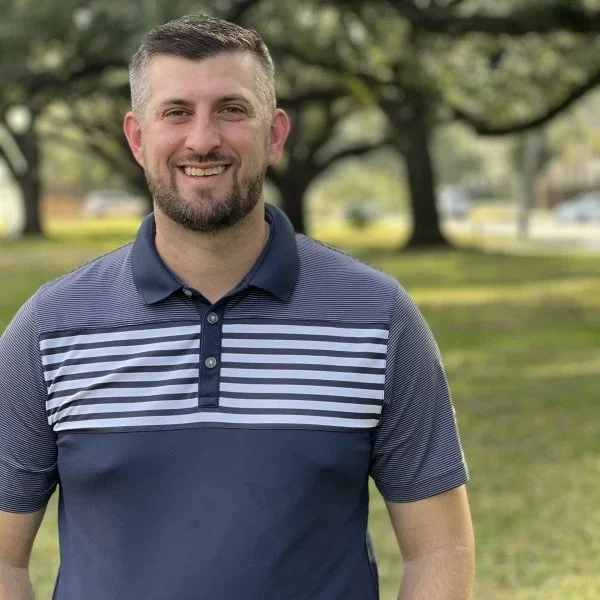 A smiling man with a beard and short hair standing outdoors in a park, wearing a navy blue and white striped polo shirt. Behind him are trees and a grassy area.