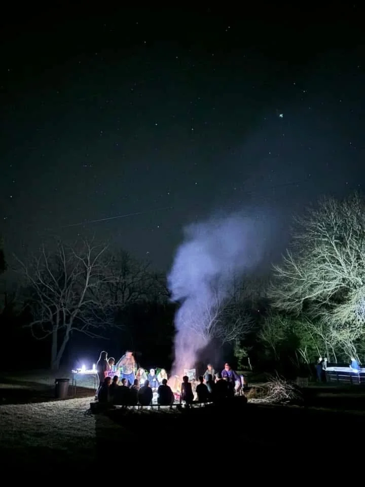 People gathered around a campfire at night, with smoke rising into the starry sky, and leafless trees in the background.