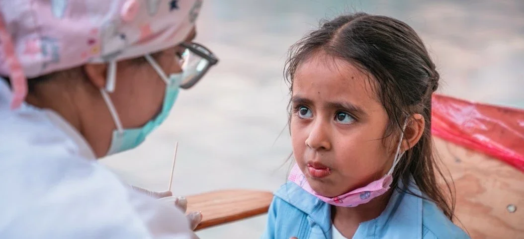 A young girl getting a COVID-19 nasal swab test from a healthcare worker in protective gear, outdoors near a body of water.