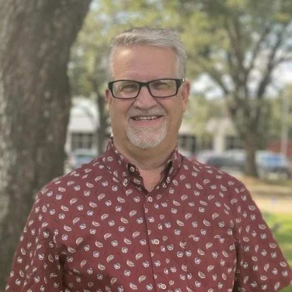 A middle-aged man with glasses and gray hair, smiling outdoors in front of a tree and residential buildings, wearing a maroon shirt with a white pattern.