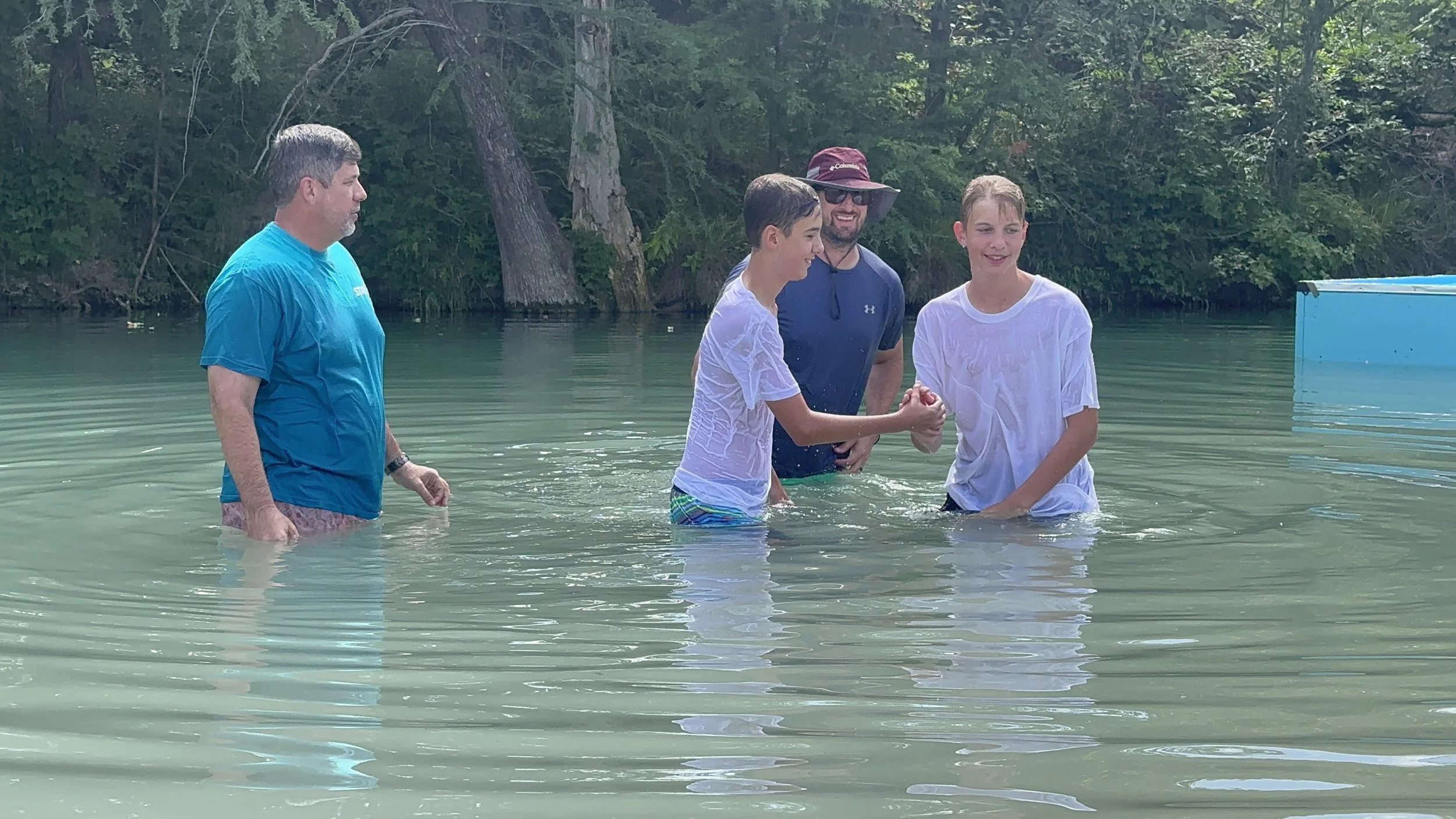 Four people in a lake, two young boys are shaking hands, surrounded by trees, with two adults watching and smiling.