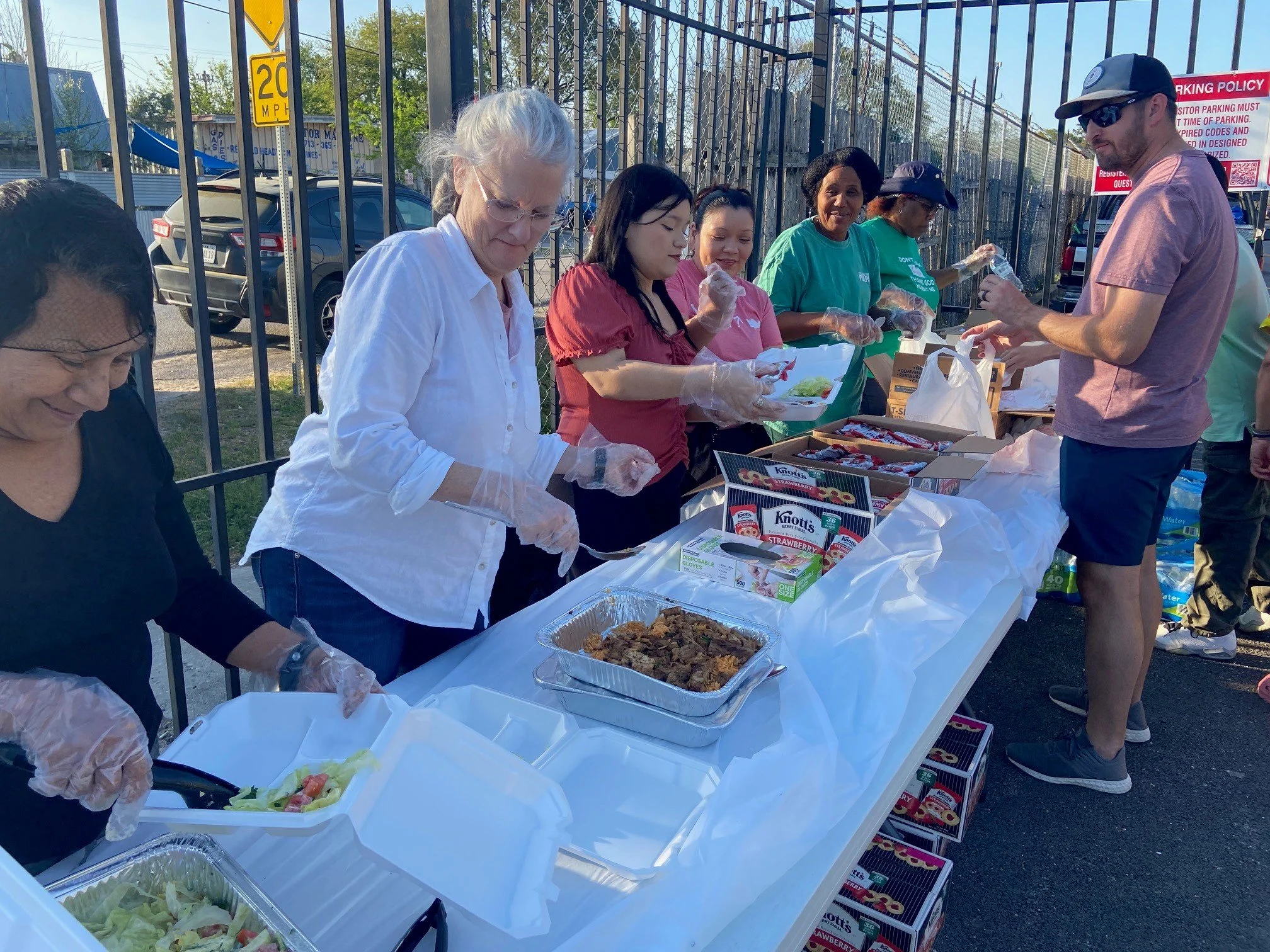 People serving and receiving food at an outdoor community event, with tables filled with trays of food, drinks, and paper plates, beside a fenced area and parked cars.