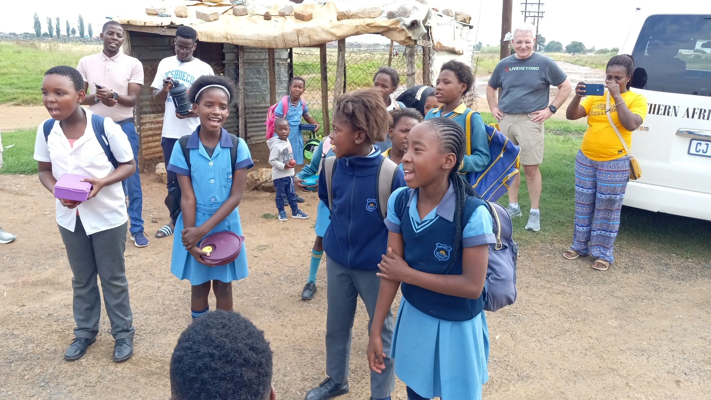 A group of children and a few adults standing outdoors in a rural area near a small building and a vehicle. The children are wearing school uniforms and smiling, some carrying backpacks and talking to each other. The adults are taking photos and observing the scene.