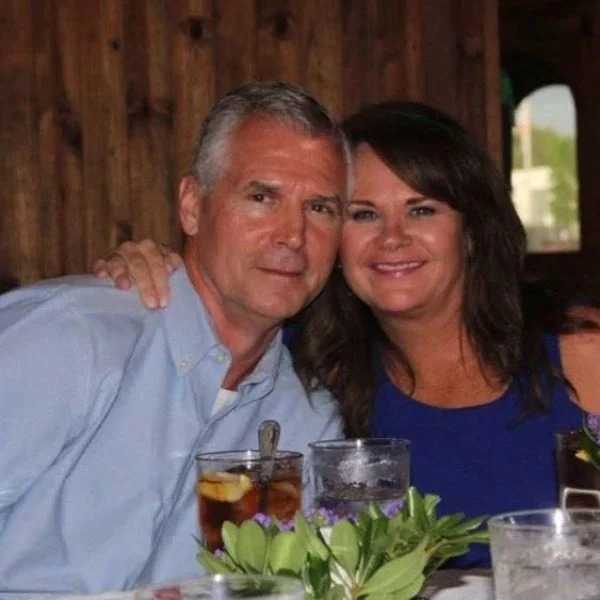 A man and woman sitting together at a table, smiling, with drinks and a floral centerpiece, inside a wooden-paneled room.