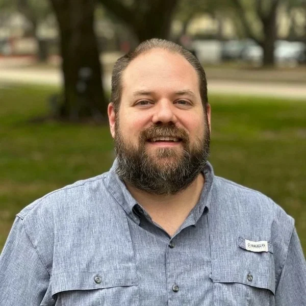 A bearded man with short hair wearing a blue button-up shirt smiling outdoors with trees and grass in the background.