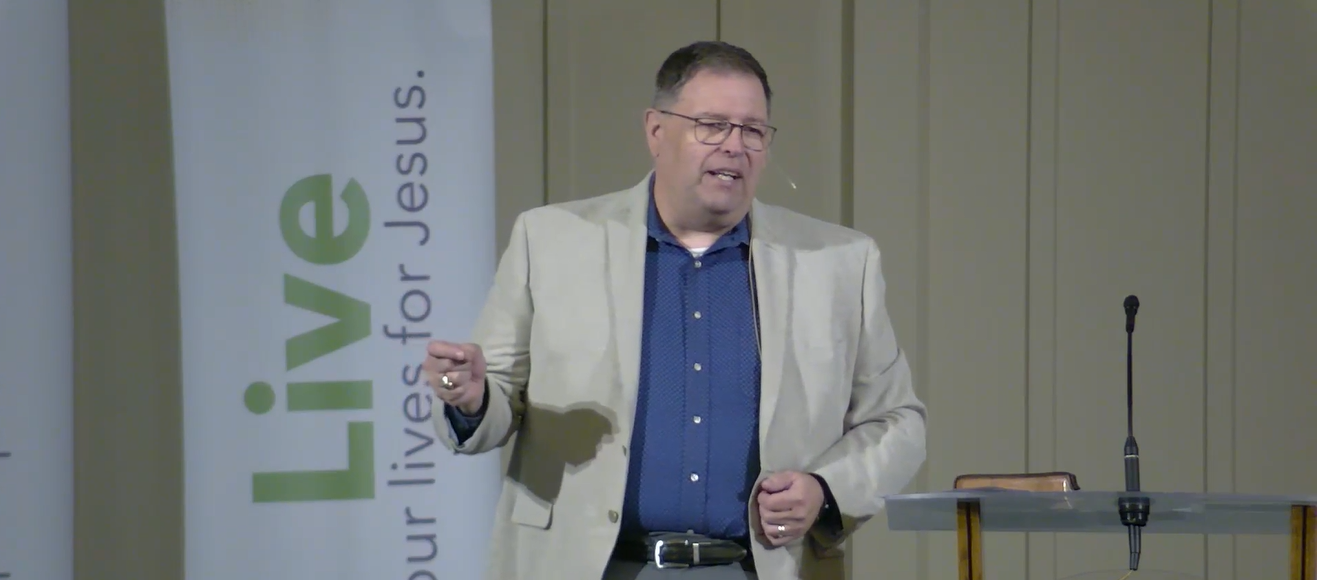 Man giving a presentation at a church or seminar, standing near a podium with a microphone, with a banner that reads 'Live: your lives for Jesus' in the background.