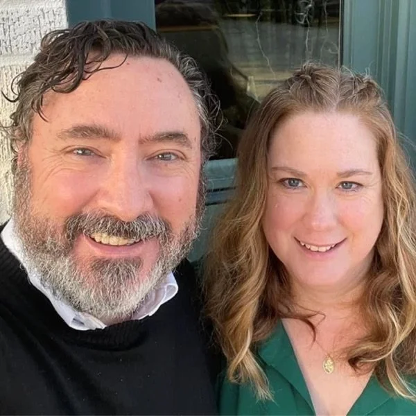 A man with curly hair and a beard and a woman with wavy hair are smiling for a selfie outside.
