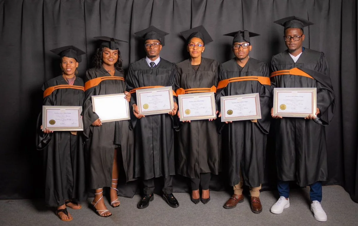 Six graduates wearing black caps and gowns with orange accents, holding diplomas, standing in front of a black curtain.