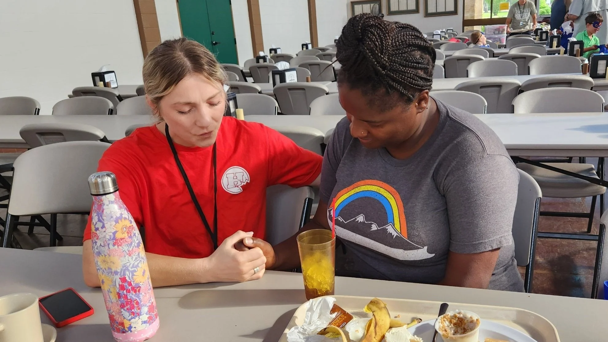 Two women sitting at a table, holding hands, with a large tray of food and drinks in front of them, in a large room with many empty chairs and tables in the background.