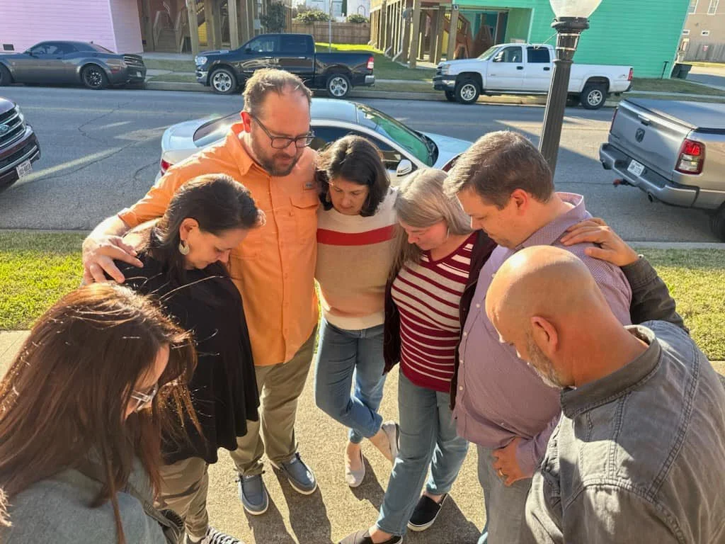 A group of nine people standing outdoors in a huddle with their heads bent down, some with arms around each other's shoulders, appearing to pray or have a moment of reflection on a sidewalk near parked cars and a lamppost.