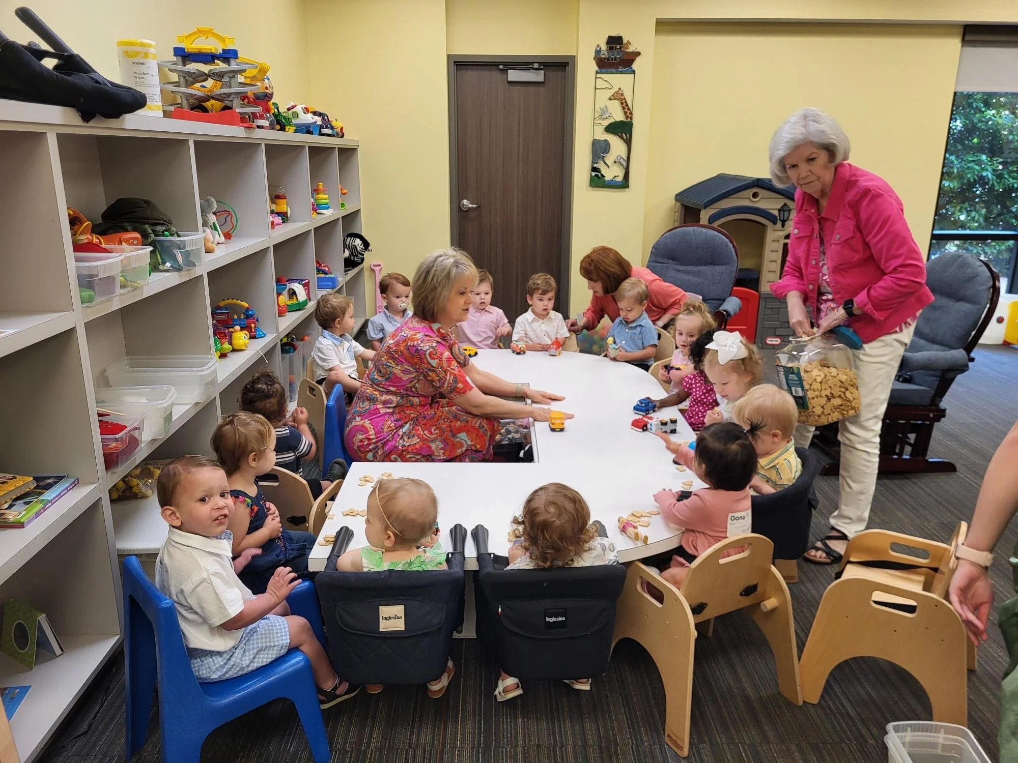 A classroom with young children and two adult women engaging in activities around a large white table. Children are seated in chairs or car seats, and the room has shelves with toys and books. One woman in a pink jacket is helping children, while the other woman in a colorful dress is seated at the table with children.