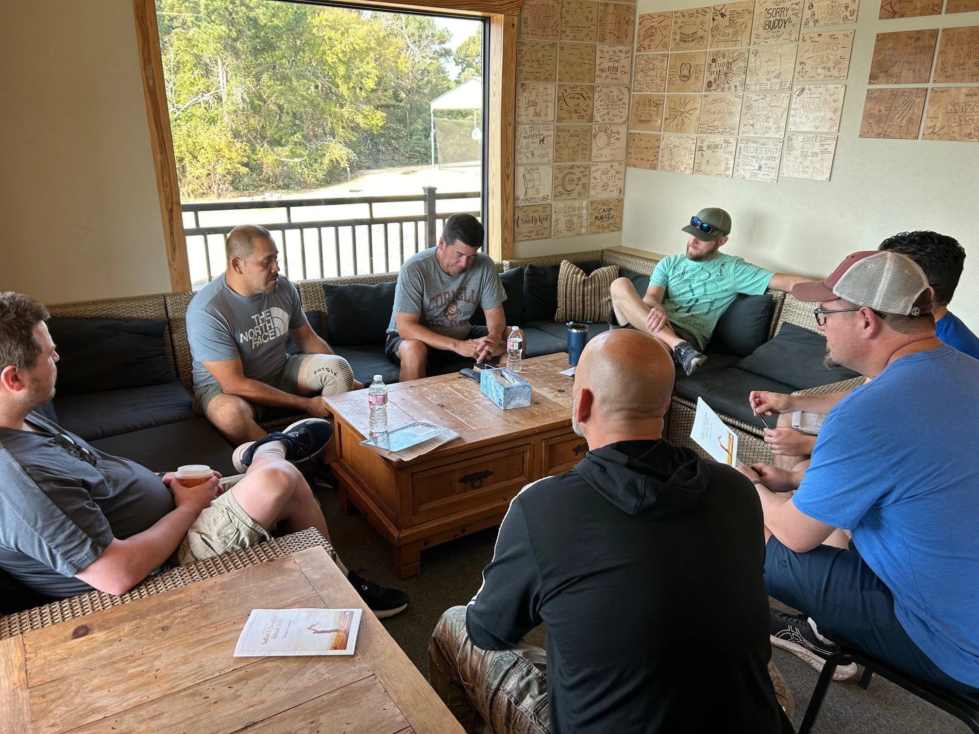 A group of seven men sitting around on couches and chairs in a semi-circle in a room with one large window and a wall decorated with many small framed artworks or prints. Some water bottles and paper products are on the table in the center.