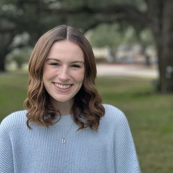 A young woman with wavy brown hair smiling outdoors in a park, wearing a light blue sweater and a necklace with a letter 'D'.