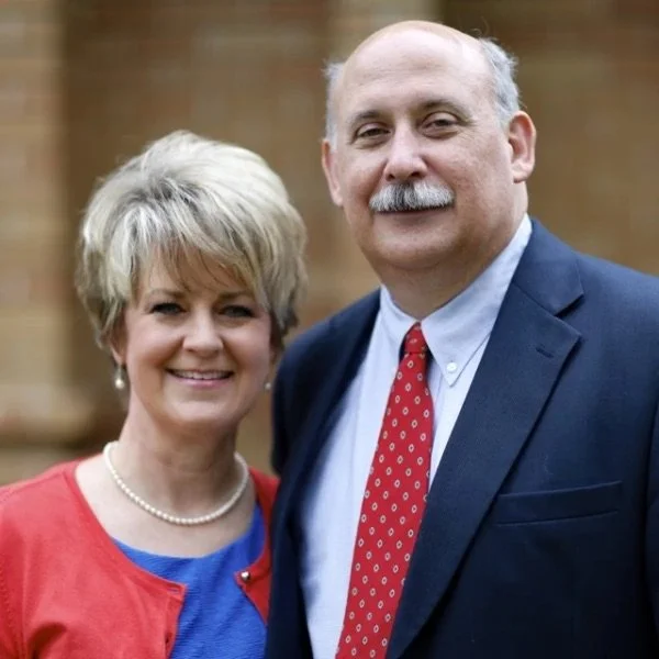 A smiling woman with short blonde hair wearing a red cardigan and pearl necklace standing next to a smiling man with a mustache, balding head, wearing a dark suit, light blue shirt, and red polka dot tie, outdoors with a brick wall background.
