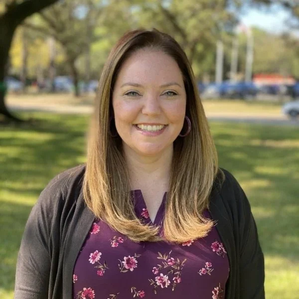 A woman with shoulder-length light brown hair, wearing a purple floral top and a black cardigan, smiling outdoors with trees and a park in the background.