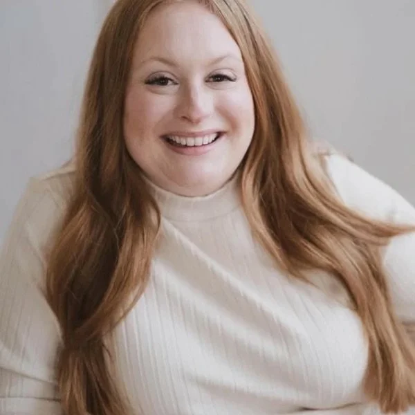 A woman with long red hair smiling in a cream-colored turtleneck shirt.
