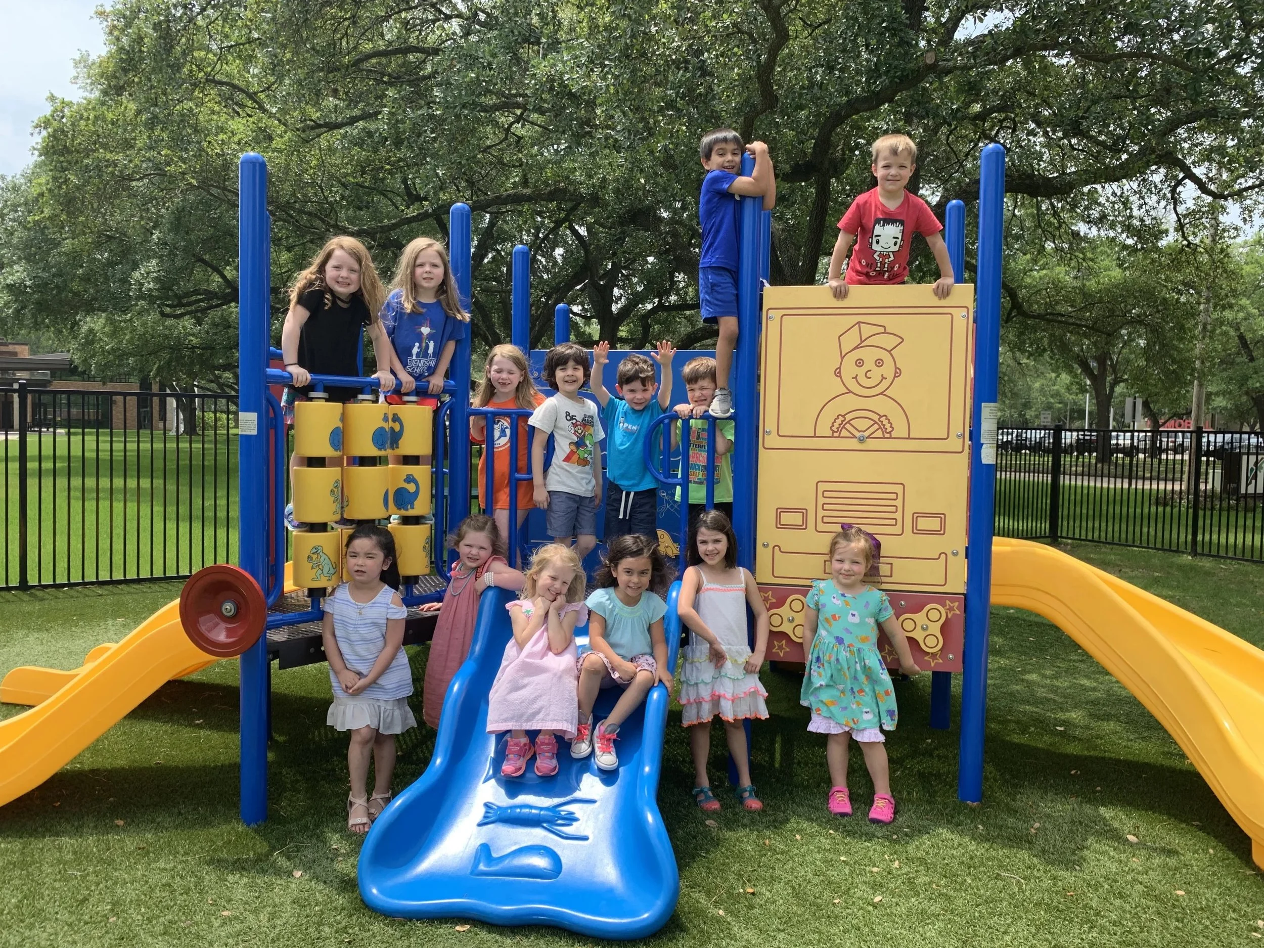 Group of children playing on a colorful playground structure with slides and climbing areas, surrounded by a black metal fence and green trees.