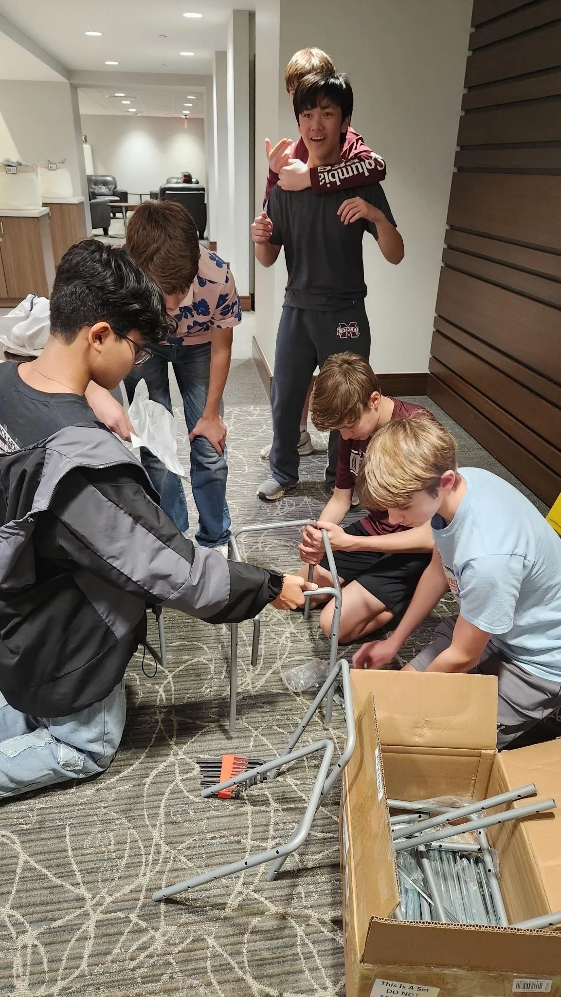 A group of children and teenagers assembling a metal wheelchair on a carpeted floor in a lobby or common area, with boxes of parts nearby.