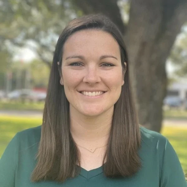 A young woman with straight brown hair wearing a green top, smiling outdoors near a tree in a park.