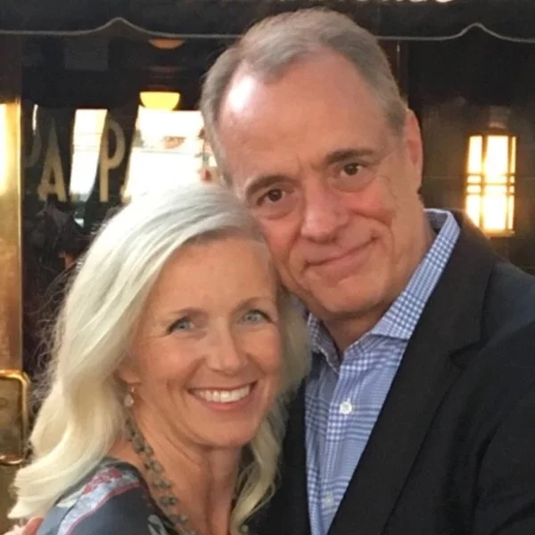 A smiling woman with long blonde hair and a man with short hair taking a selfie together in a dimly lit indoor setting.