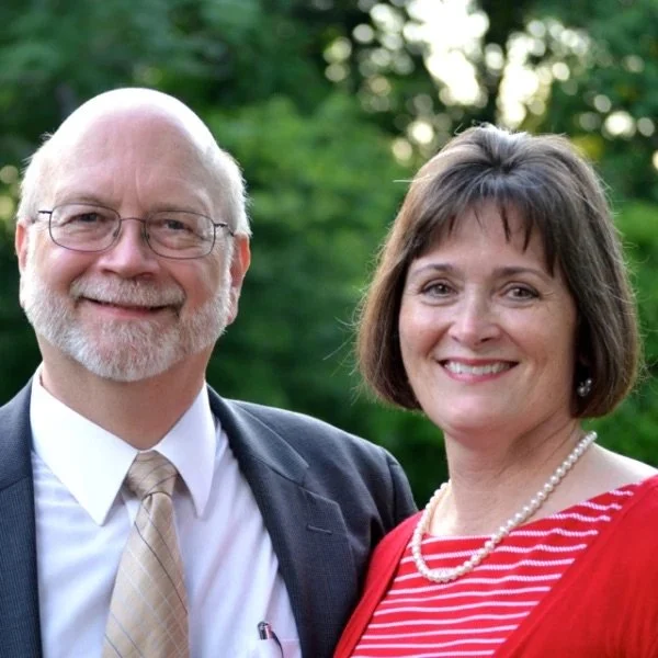 A smiling older man with glasses, a gray beard, and white hair, dressed in a suit and tie, standing next to a smiling woman with short brown hair, wearing a red and white striped blouse and pearl necklace, outdoors with green trees in the background.