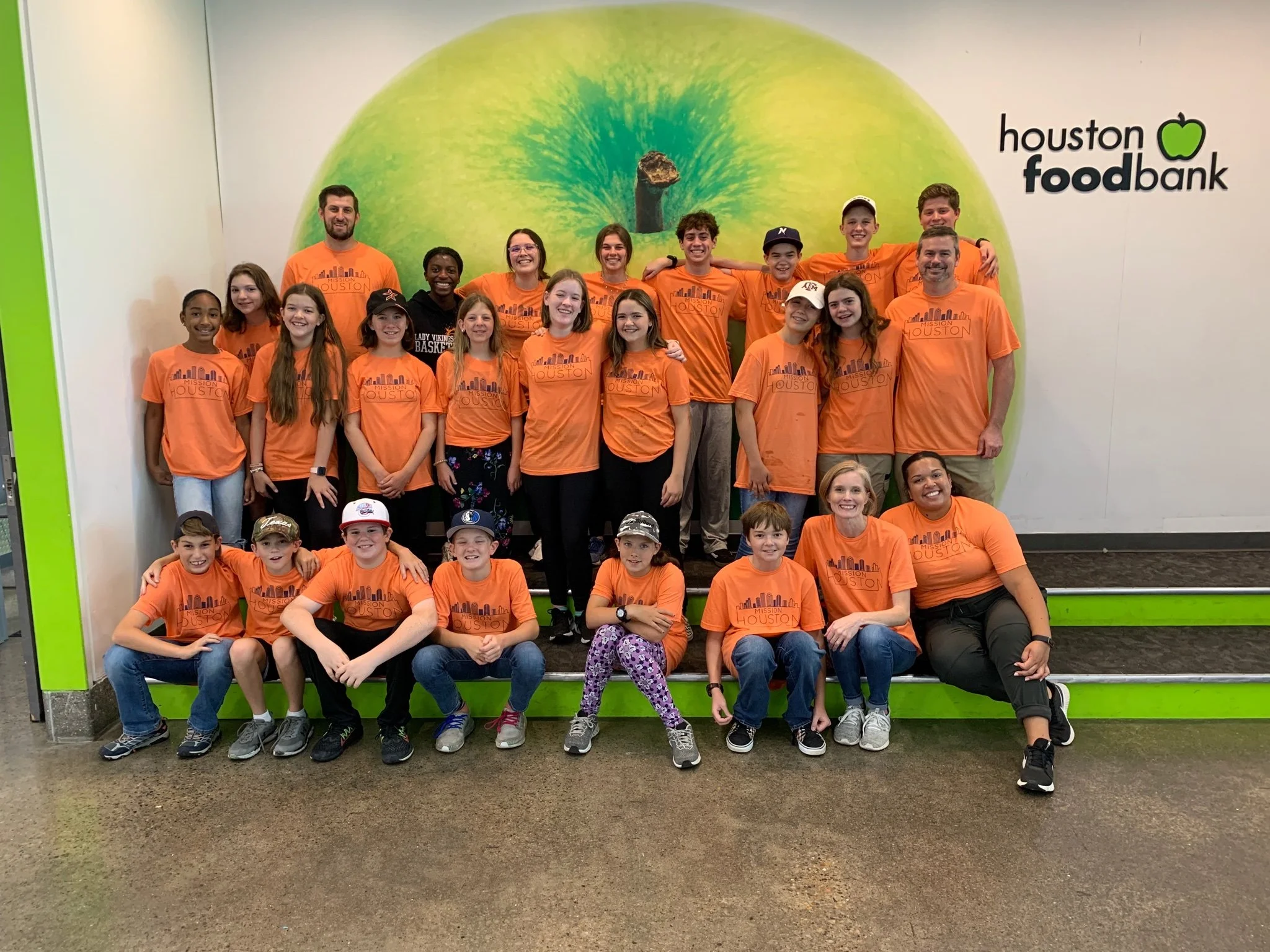 Group of children and adults wearing orange t-shirts with Houston Food Bank logo, posing for a photo indoors in front of a large green apple graphic on the wall.