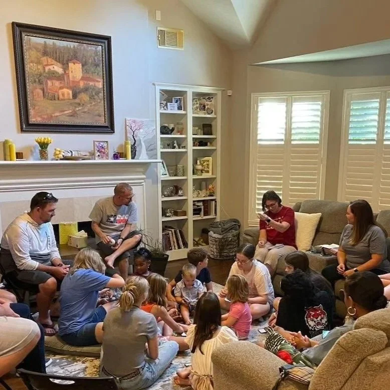A group of children and adults gathered in a living room, sitting on the floor and couches, engaging in a group activity or story time near a fireplace with decorative items on the mantel, a tall shelf with books and decorations, and windows with plantation shutters.